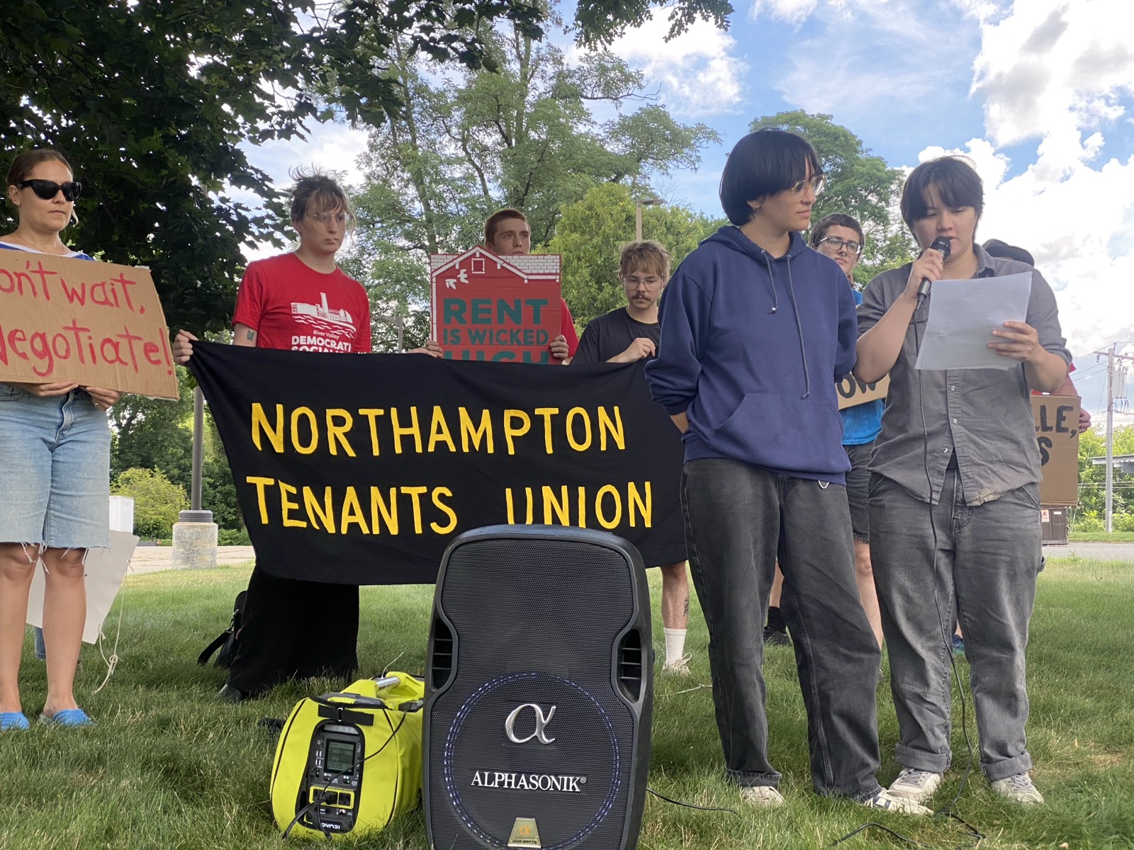 Maia Penuel and Milla Wenick, tenants of 126 King St. in Northampton, speak at a rally on July 10, 2026. The two speak into a microphone on the right side of the picture, a large speaker sitting to their left. Behind them, several people hold a black banner that reads, in yellow font, "Northampton Tenants Union." Others hold signs saying "Don't wait, negotiate" and "Rent is Wicked High."
