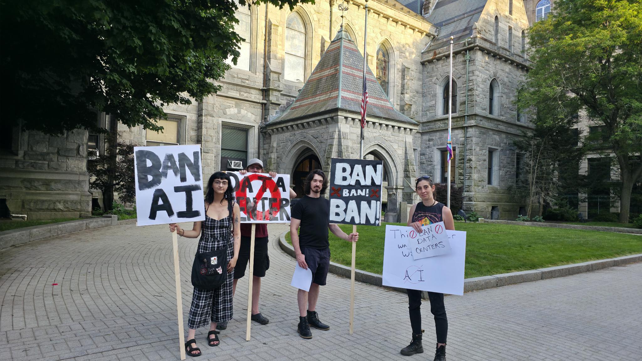 Protesters stand outside Holyoke City Hall on June 11, 2026, with signs in protest of AI data centers. They read: "Ban AI," "Ban! Ban! Ban!", "Ban Data Ceners." One says "Data center," which is crossed out in red ink.