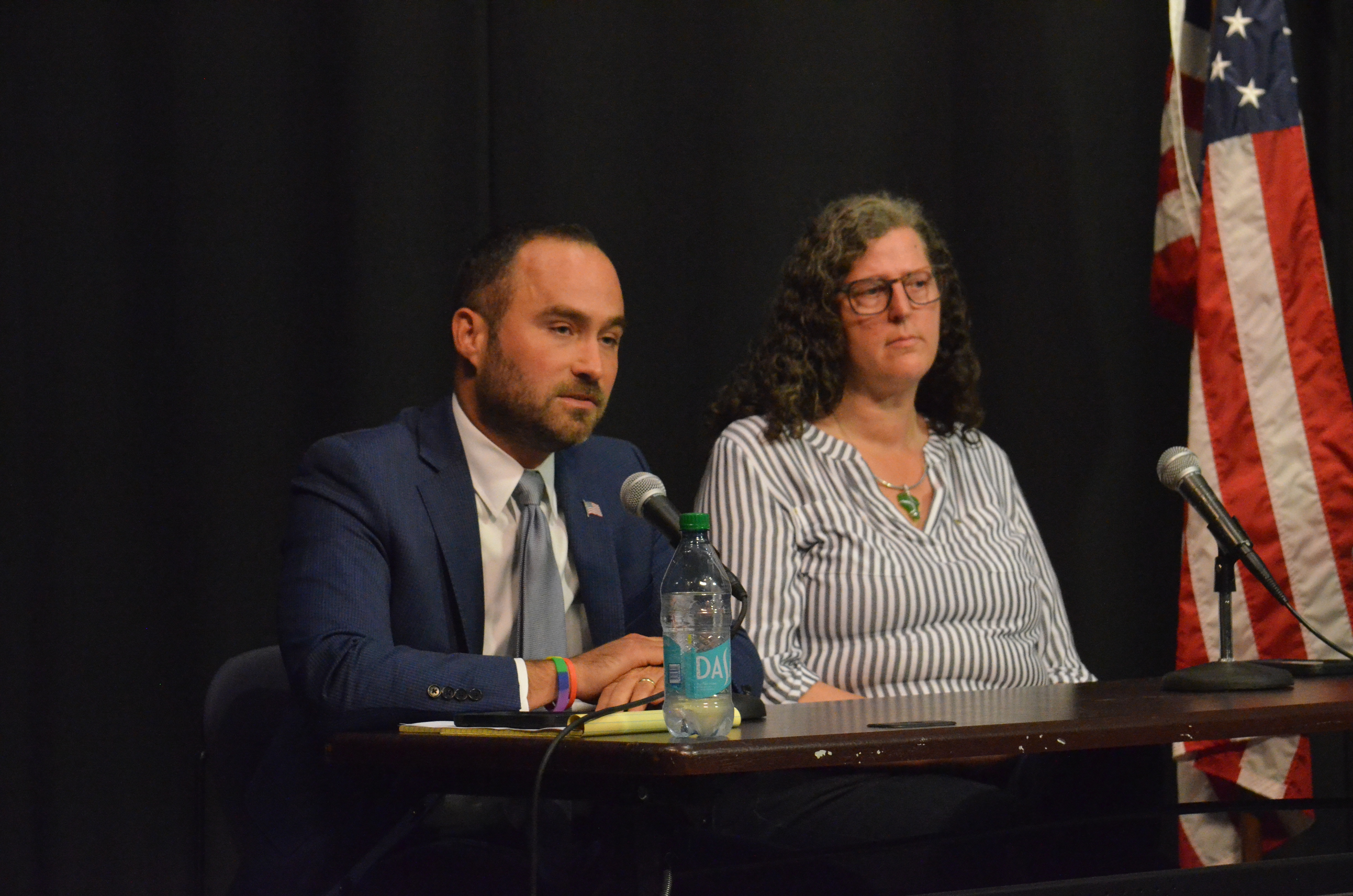 Michael Fenton, leaning forward to speak, and Tara Jacobs, are seated against a black background and an American flag.
