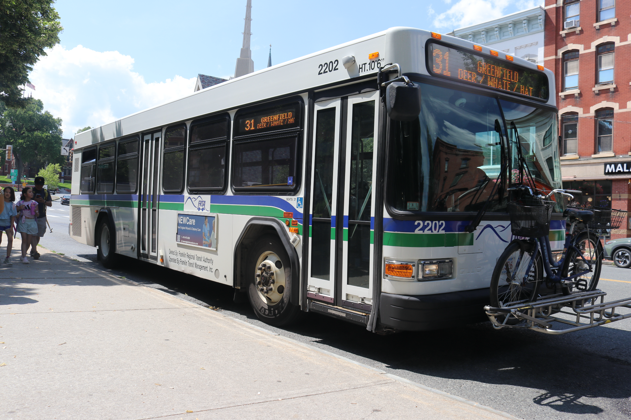 A transit bus angles towards the viewer with a bus on its front rack on a sunny day. Pedestrians are passing by and apartment buildings are visible in the background.