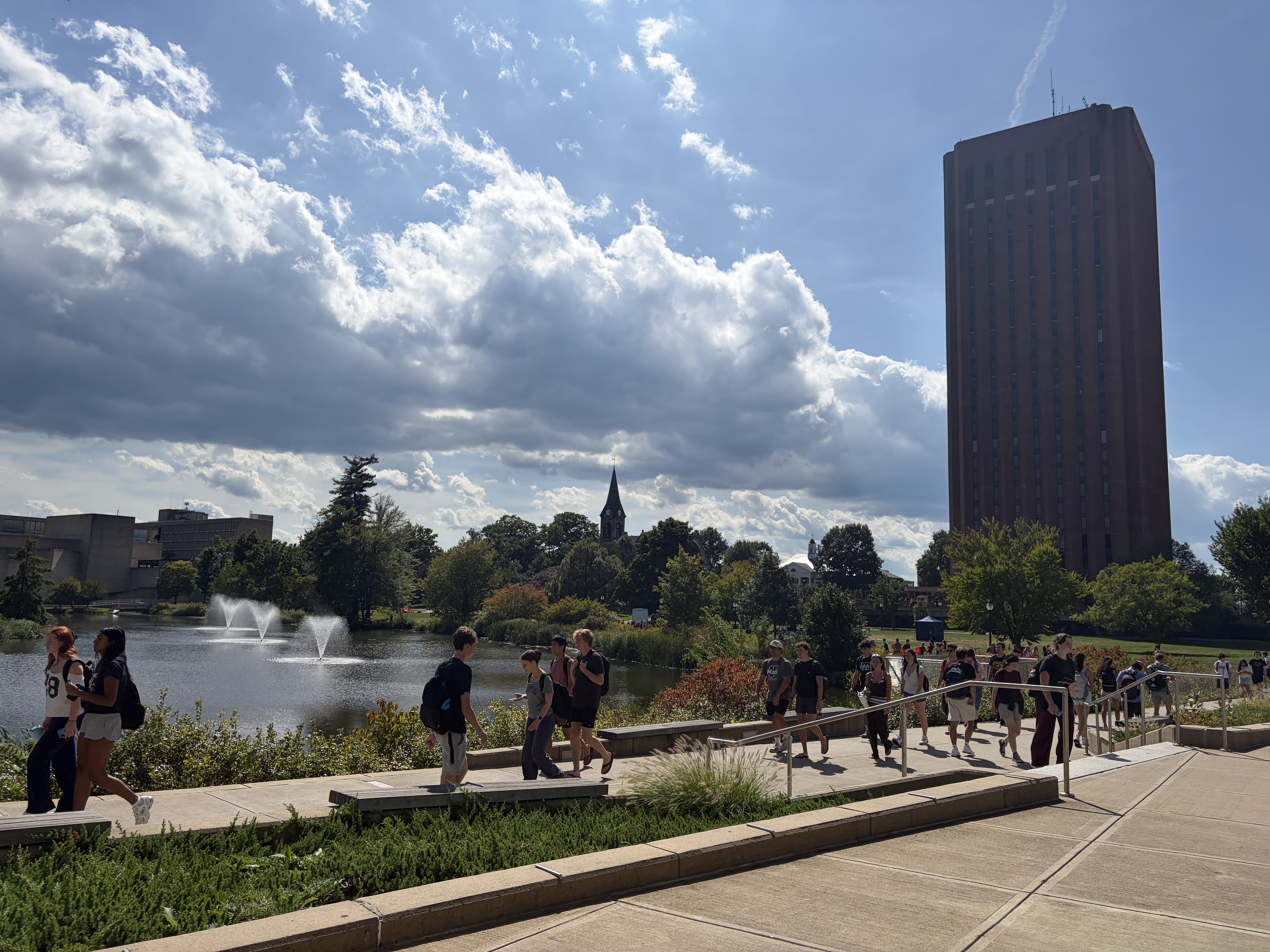 Students walk through the University of Massachusetts Amherst campus on Sept. 10, 2026. The campus pond is visible on the left, with three fountains spraying water into the air. On the right, the W.E.B. Du Bois Library towers over campus. Students are walking in the foreground.